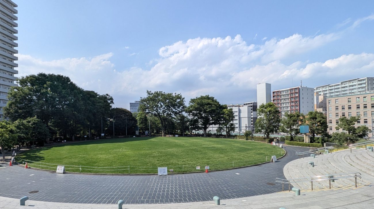 芝生の円形広場と木立、奥に並ぶ駅前の高層ビル(埼玉県川口市、2025年撮影)